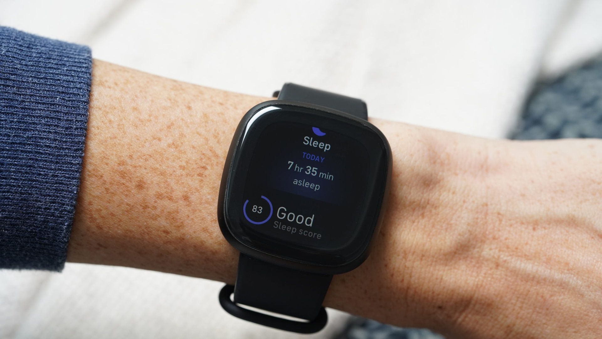 A Fitbit Versa 3 on a user's wrist displays their total sleep time and a sleep score.
