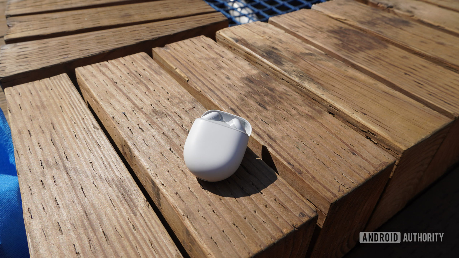 The Pixel Buds A-Series earbuds in their case sitting on a wooden bench outdoors.