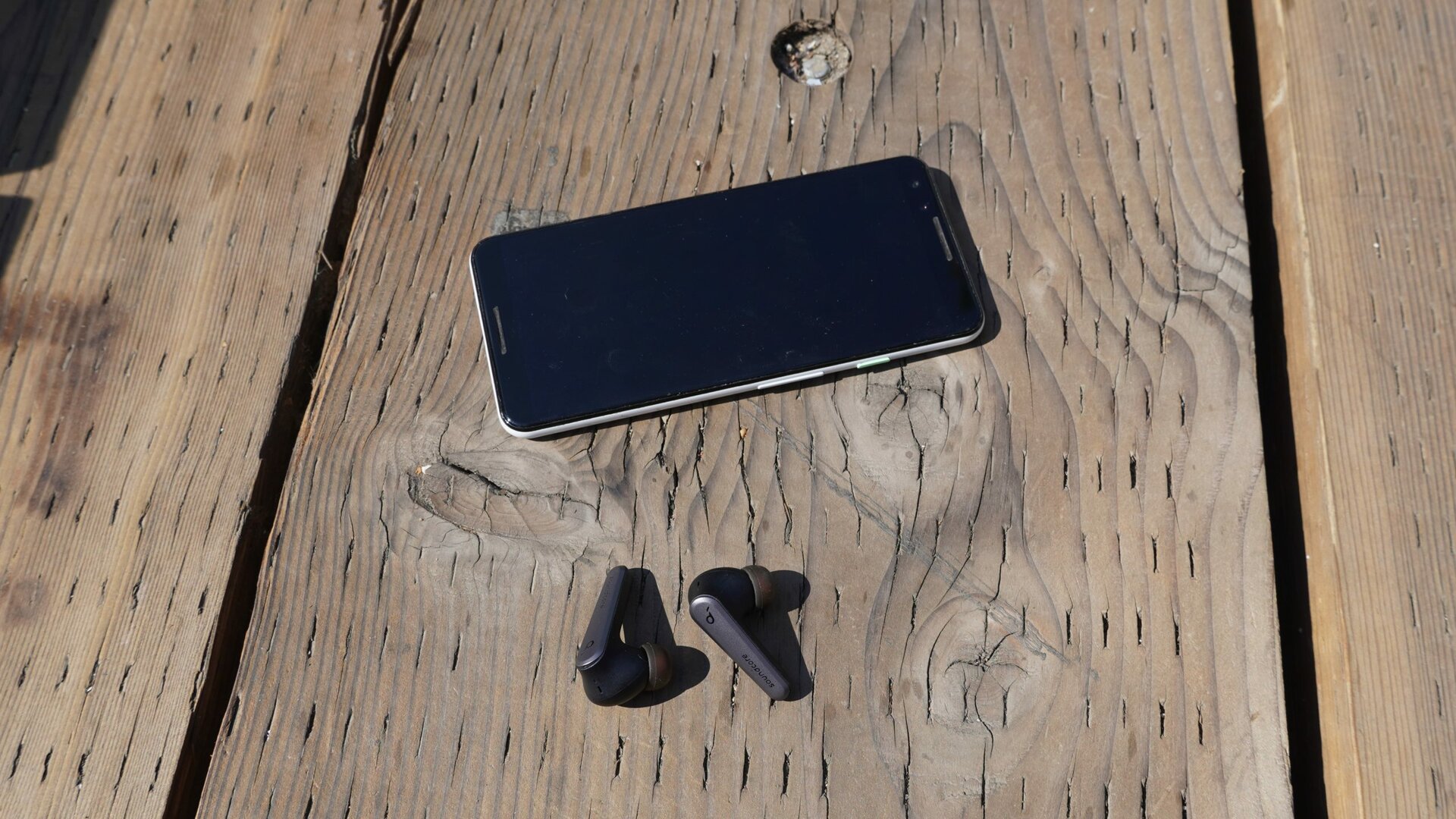The Anker Soundcore Liberty Air 2 Pro earbuds sitting next to a smartphone outdoors on a wood surface.