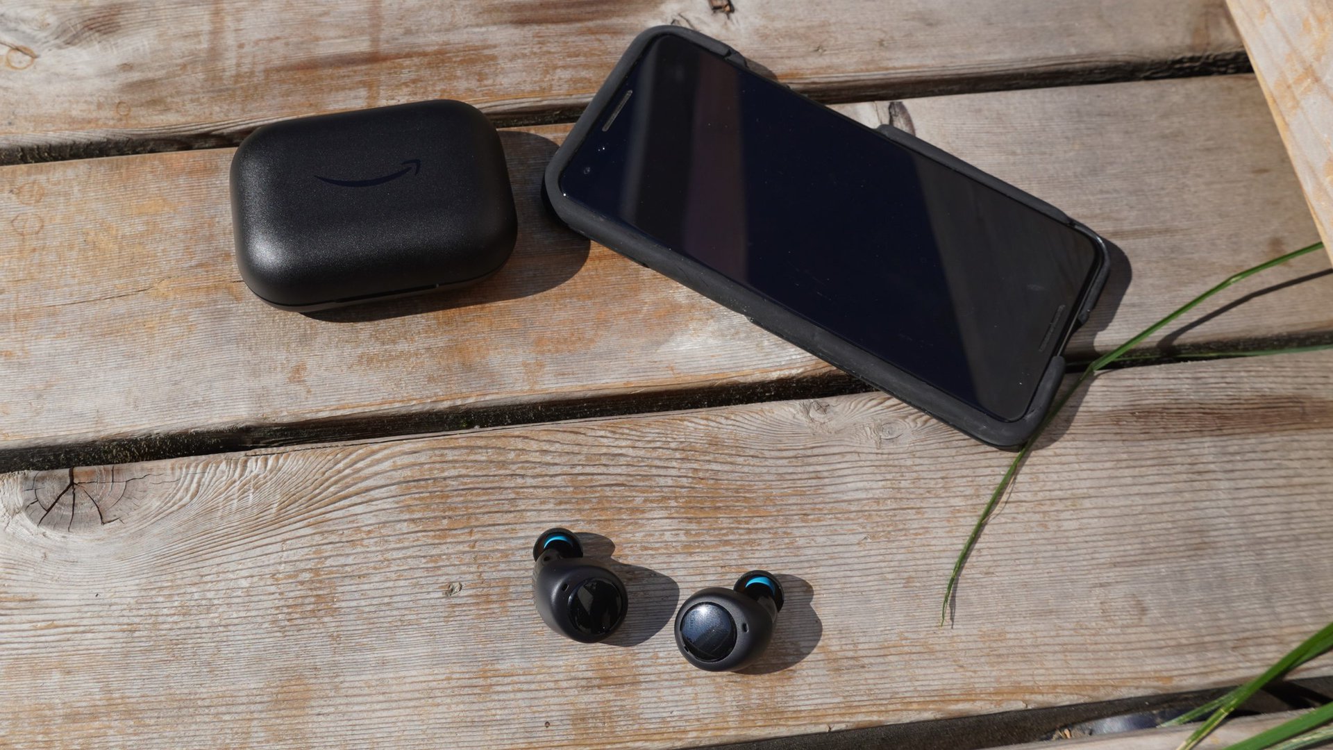 A pair of Amazon Echo Buds (2nd Gen) sitting in front of a phone to their right and their case to their left on a wood surface outdoors.