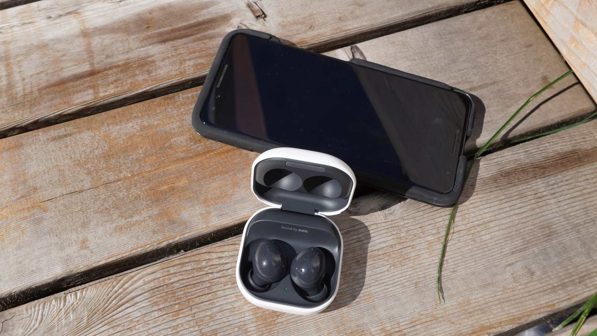 The Galaxy Buds 2 in their case sitting next to a smartphone on a wooden bench outdoors.