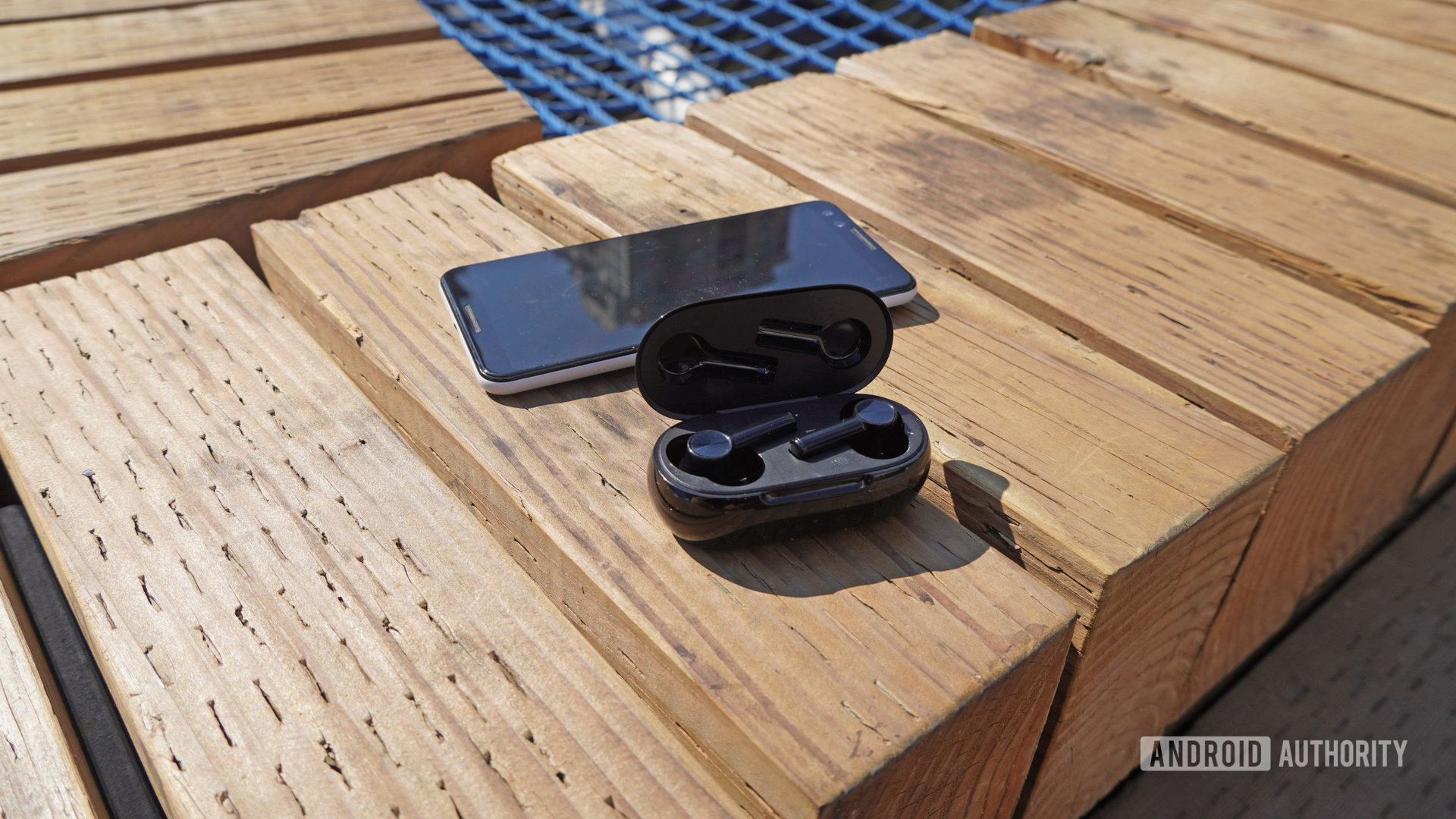 The OnePlus Buds Z2 sitting in their case on a wooden bench next to a phone.