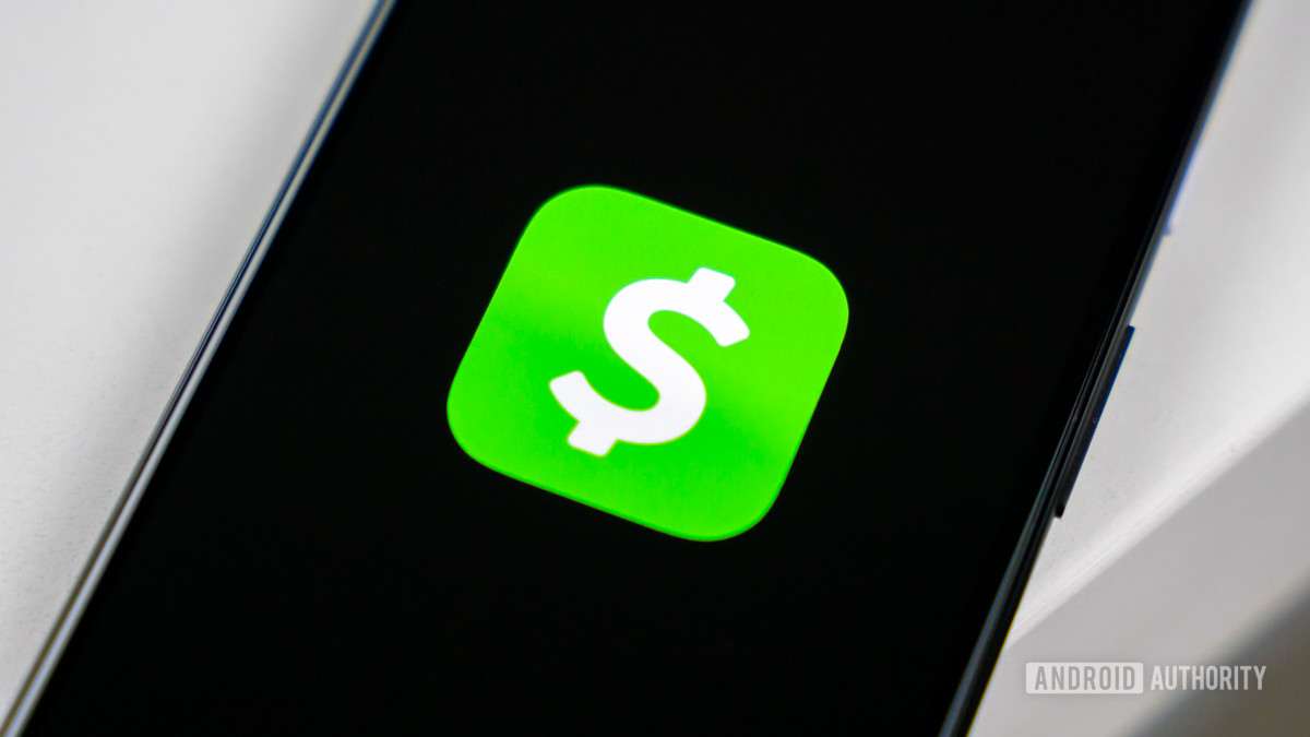 Изображение логотипа Cash App на темном экране смартфона.  Он стоит на краю белого стола.