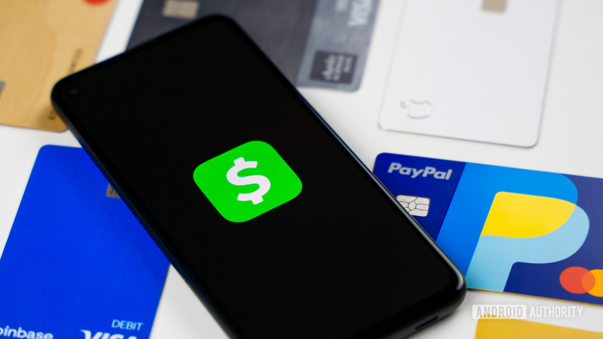 A stock photo of the Cash App logo on a dark smartphone screen. The phone rests on some credit and debit cards on white surface.