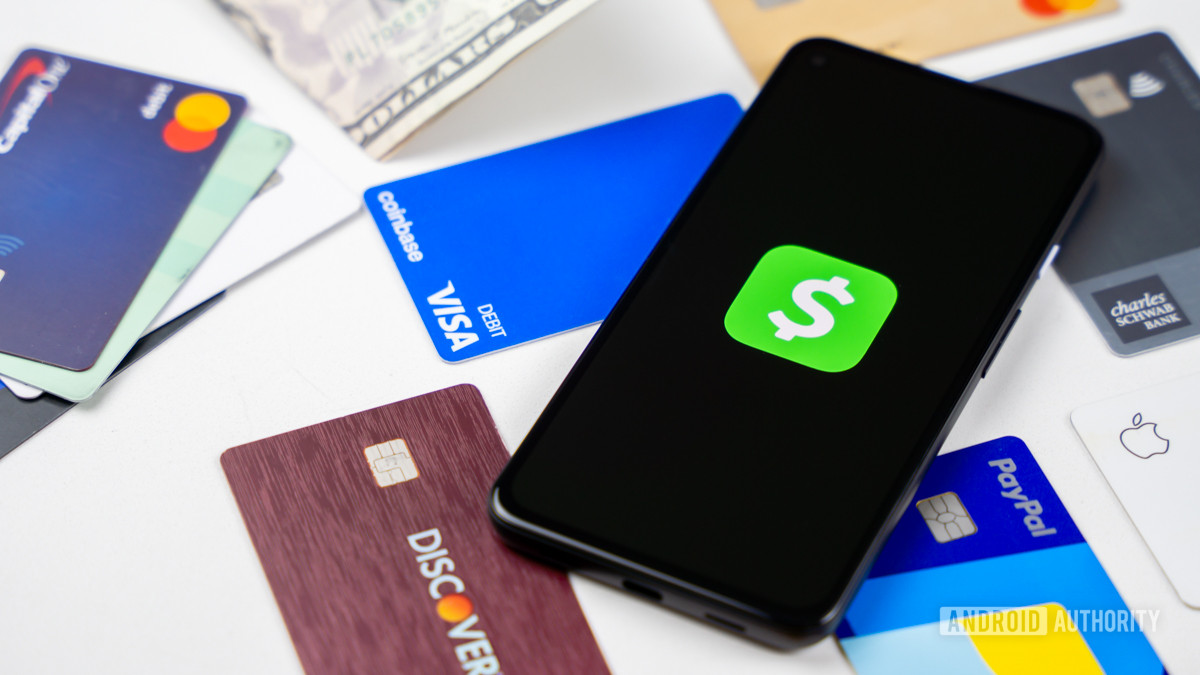 A stock photo of the Cash App logo shown on a darkened smartphone screen. The smartphone lies on top of some debit and credit cards and next to a pile of more cards and a small pile of cash.