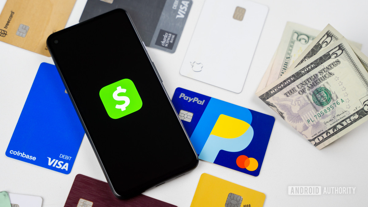 A stock photo of a smartphone with a dark screen showing the Cash App logo. It lies on some credit and debit cards next to a small pile of $5 bills.