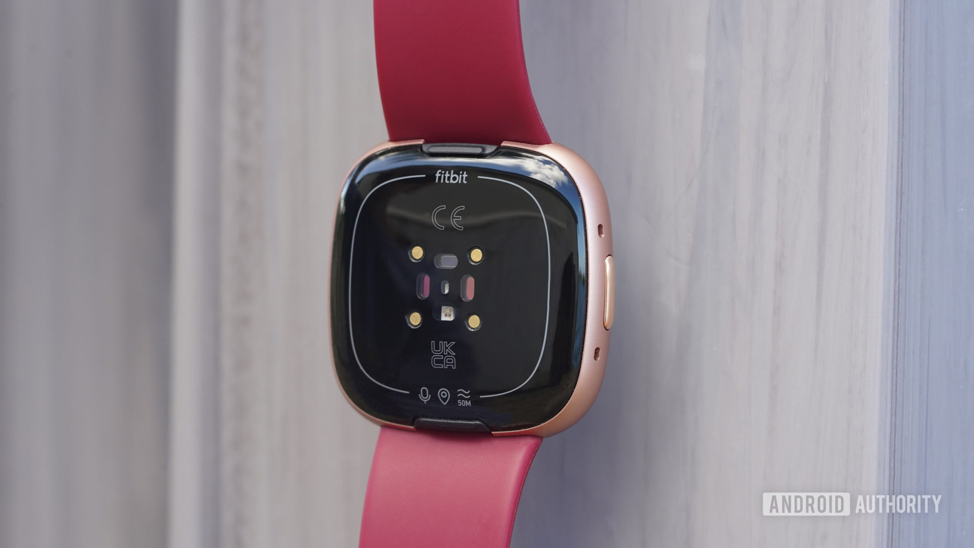 A Fitbit Versa 4 hangs in front of a gray surface, displaying the device's sensors.