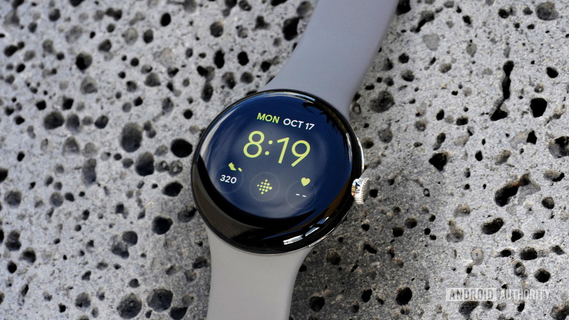 A Google Pixel Watch rests on a lava rock displaying the Utility watch face with a Fitbit shortcut.