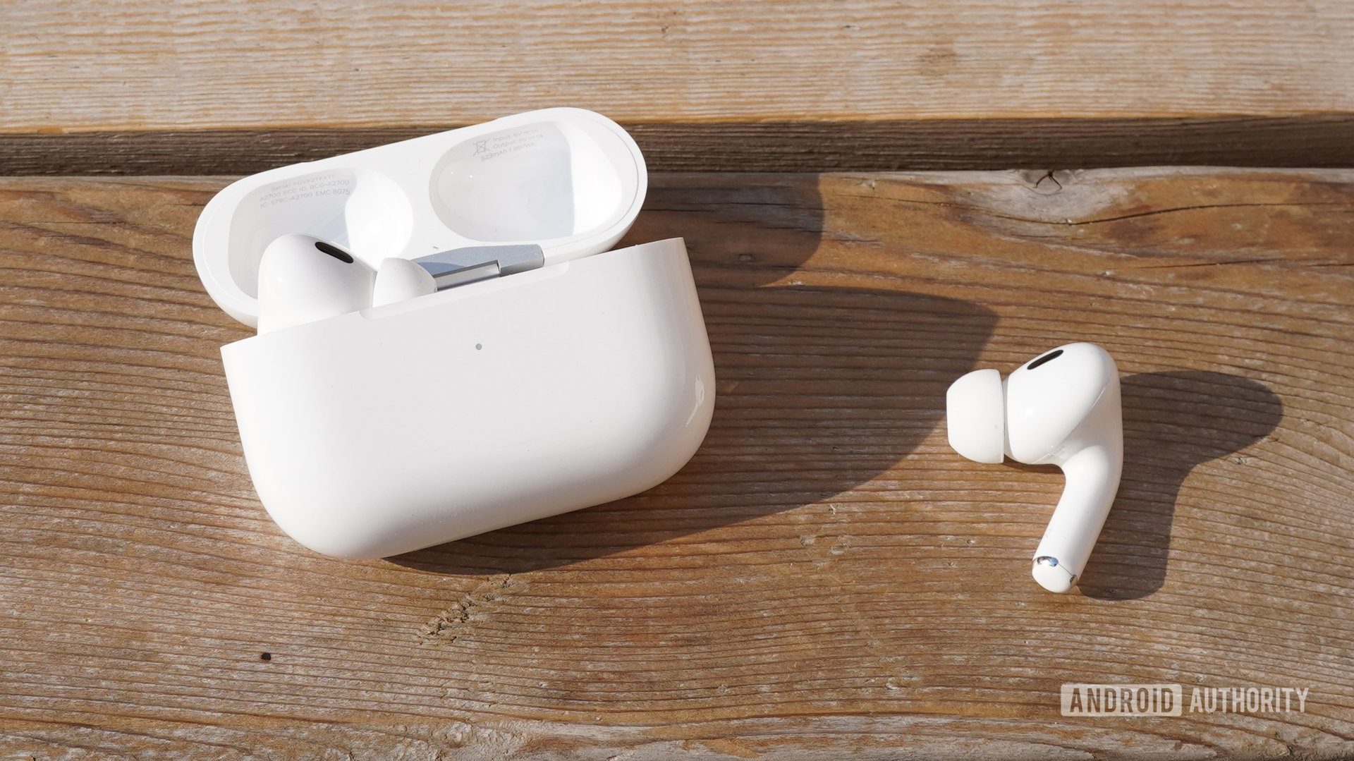 The AirPods Pro 2 with one earbud sitting outside their case on a wooden surface with the case nearby.