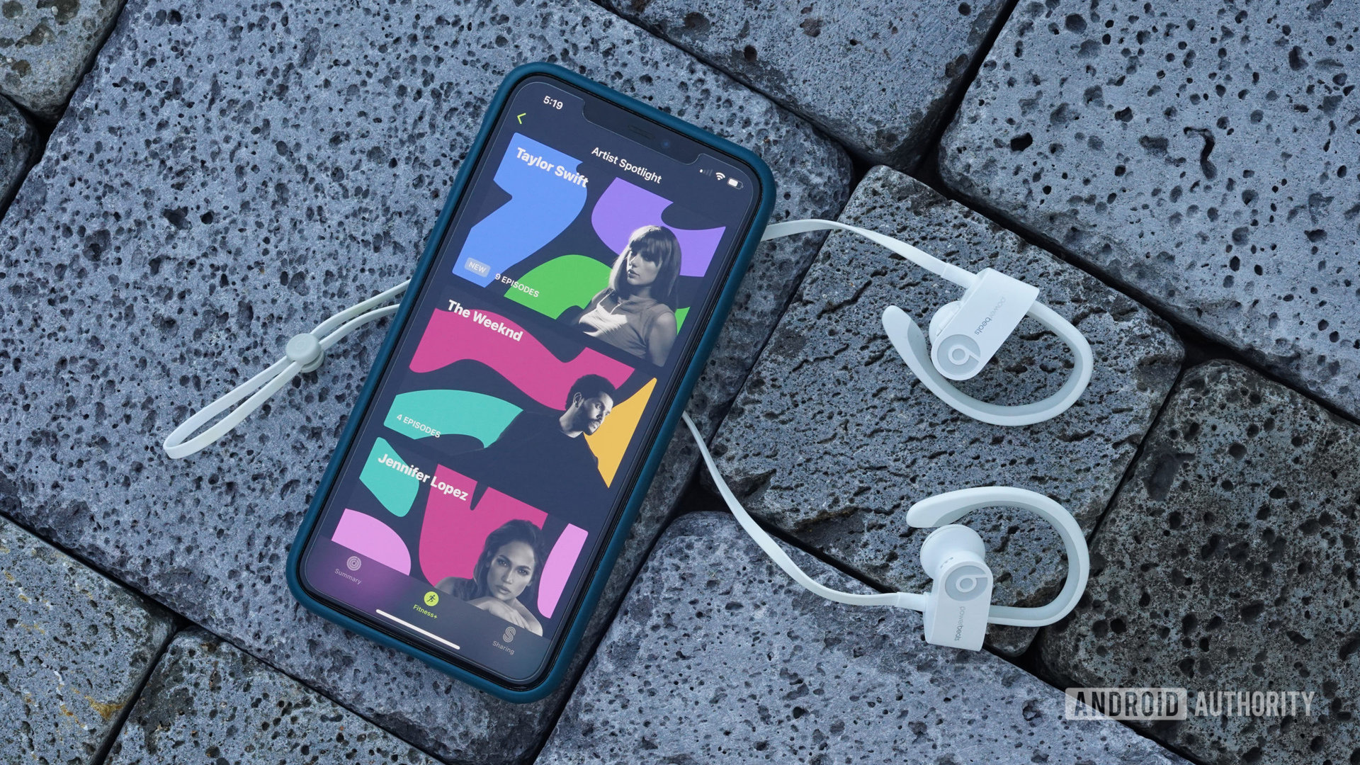 An iPhone displaying Artist Spotlight rests on a brick path along with a pair of wireless headphones.