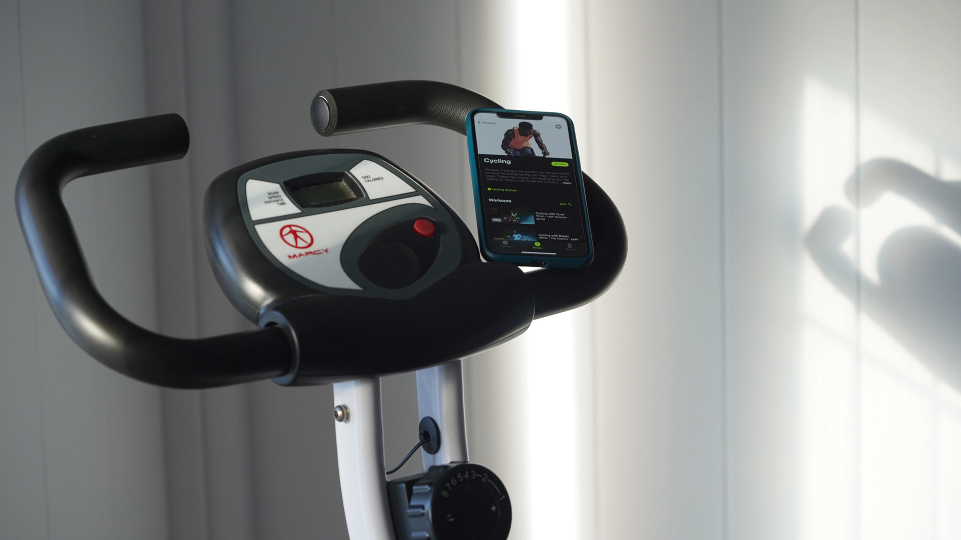An iPhone resting on a stationary bike handle displays the app's Cycling menu.