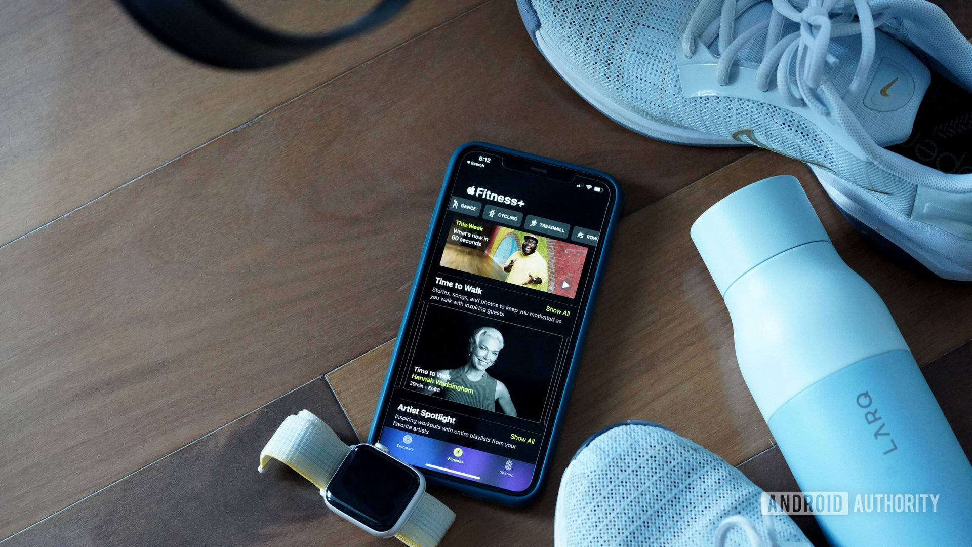 iPhone yang menampilkan Apple Fitness Plus berada di lantai studio di antara sepatu tenis, botol air, dan Apple Watch SE 2.