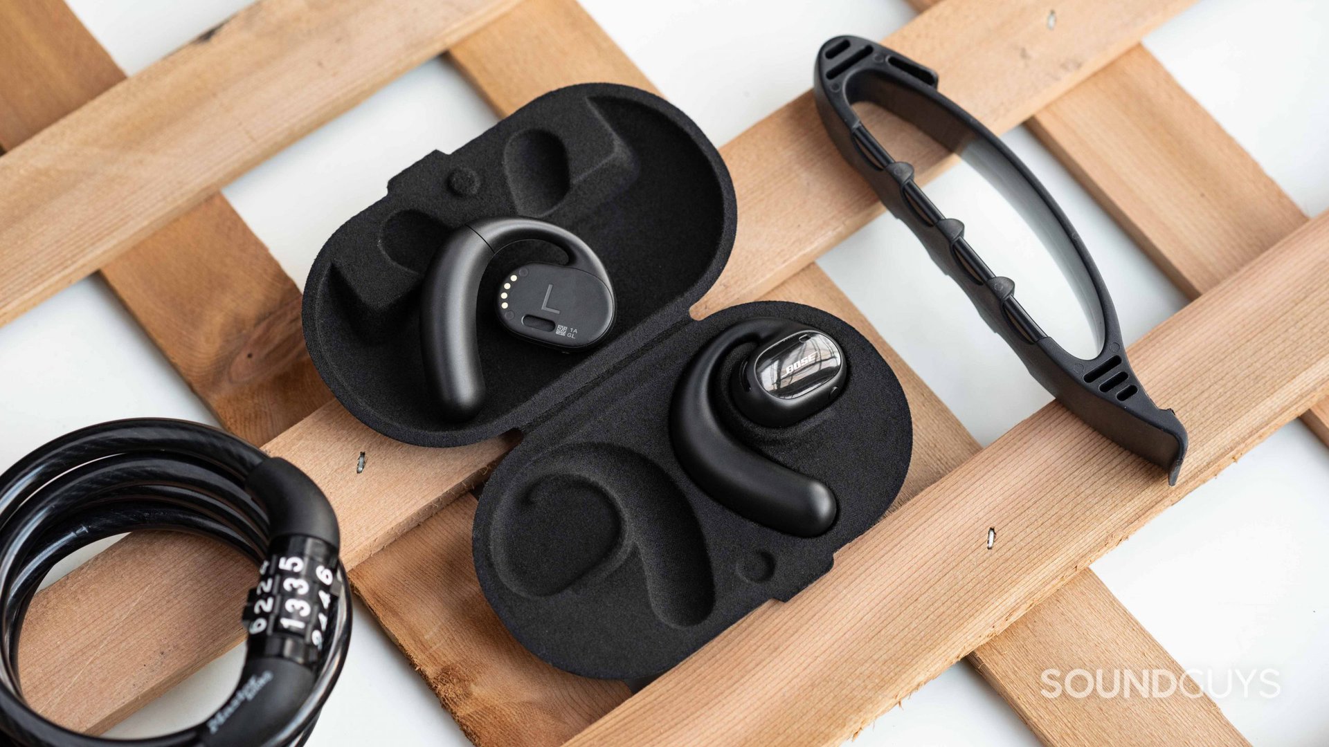 Bose Sport Open Earbuds sitting inside their case on a wooden lattice next to a bike lock and a fitness band with one earbud lying in the lid and the other in its resting spot.