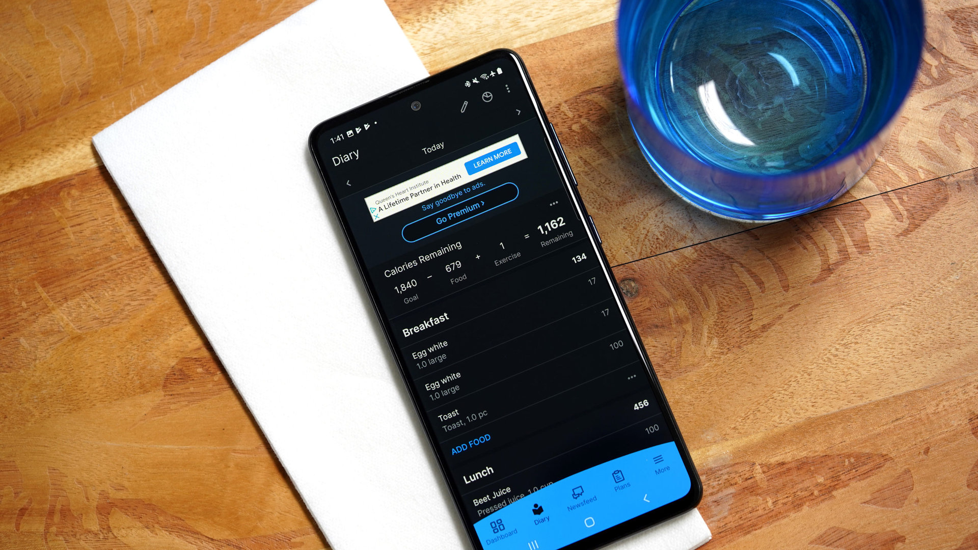 A Galaxy A51 resting on a kitchen table displays a user's food diary in the MyFitnessPal app.
