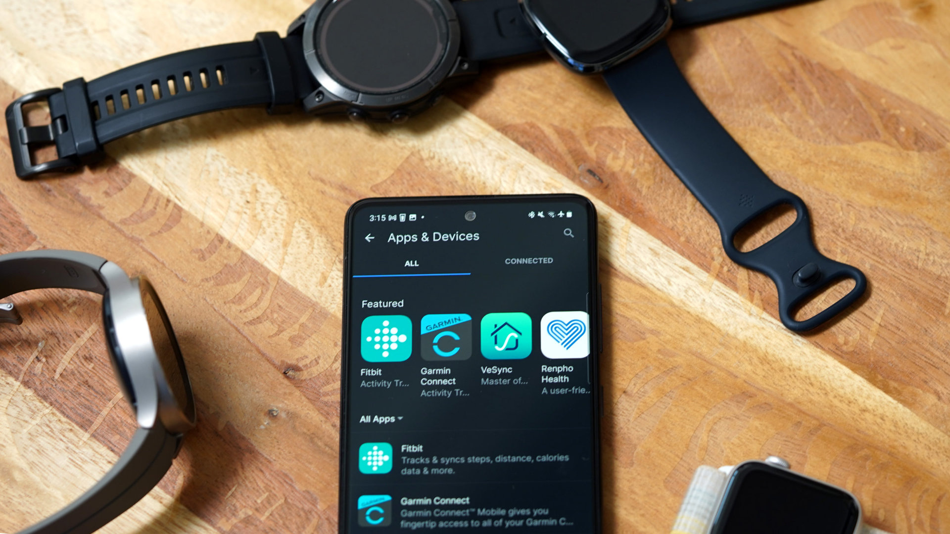 A Galaxy A51 displaying the MyFitnessPal Apps &amp; Devices screen rests on a wooden table alongside a variety of wearables.