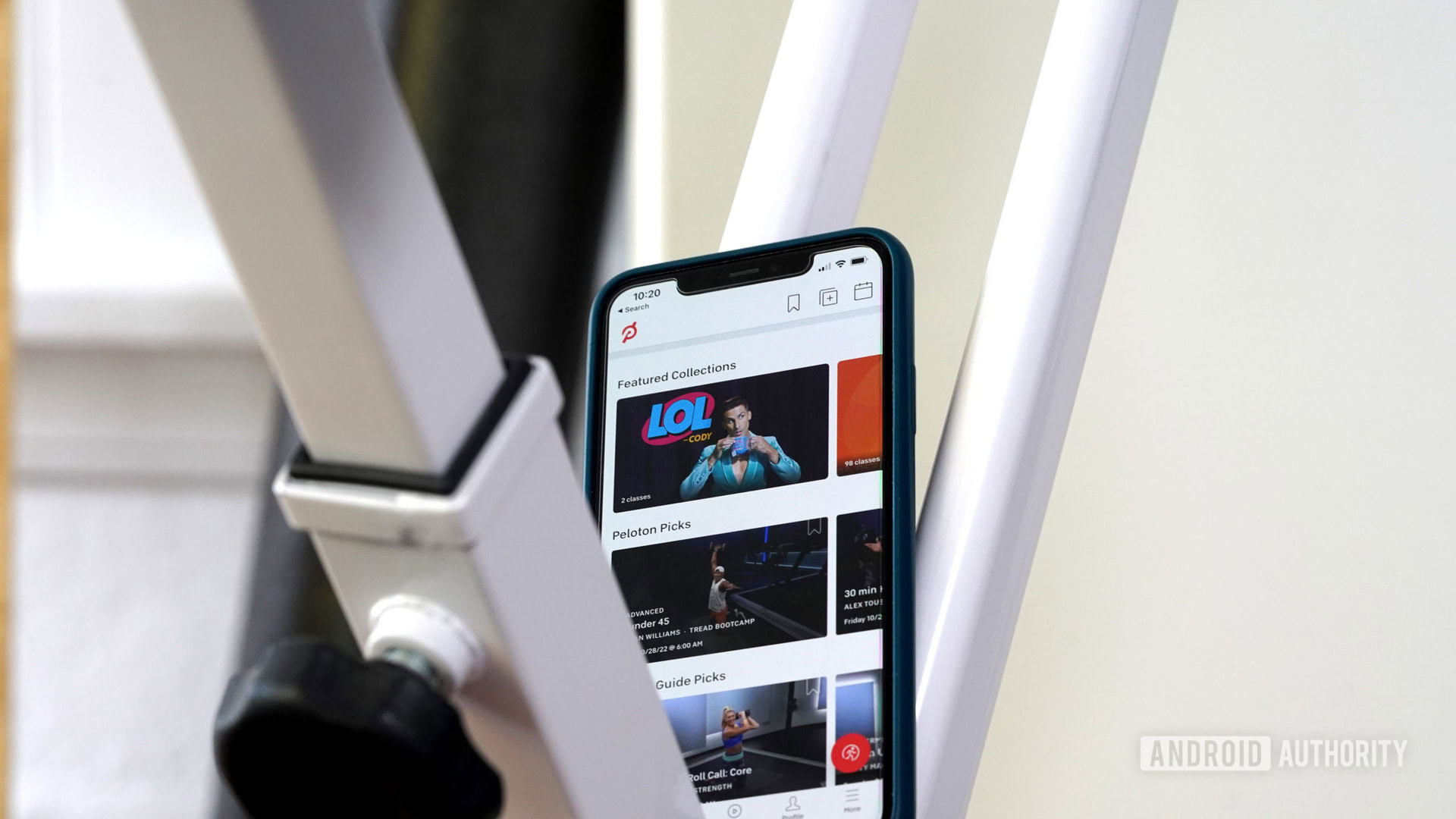 An iPhone balancing on an exercise bike displays the home screen of a user's Peloton App.