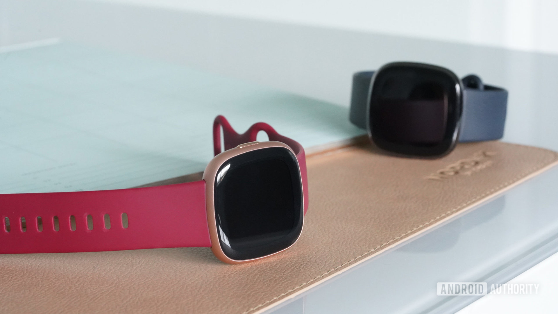 A Fitbit Versa 4 rests on a desk next to a Fitbit Sense.