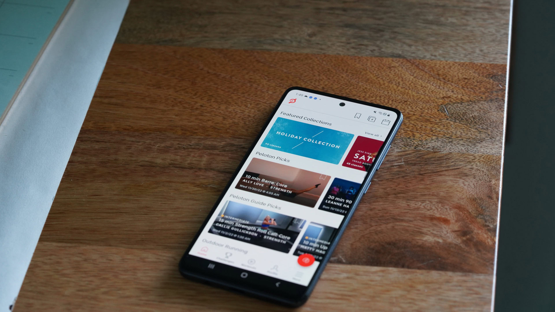 A Galaxy A51 rests on a wood surface displaying the Peloton App Home Screen.