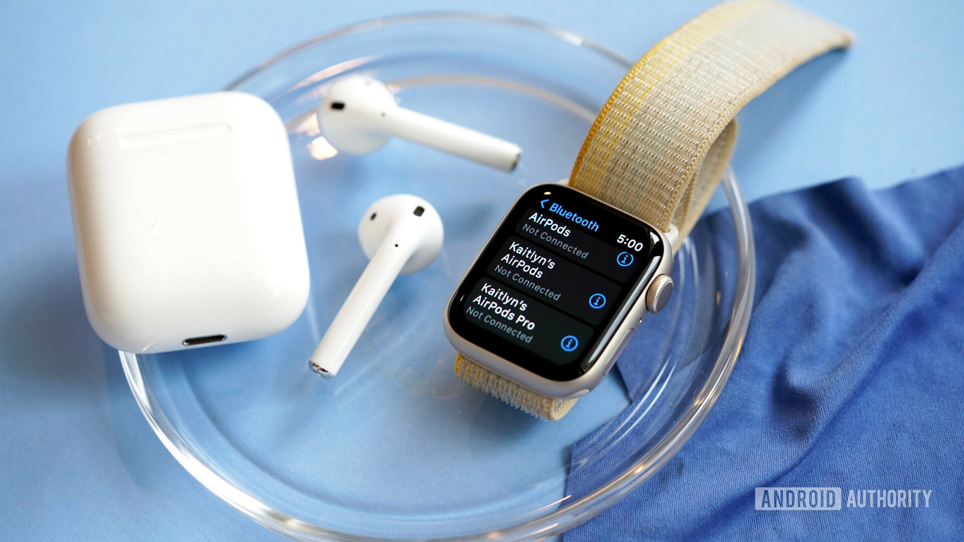 An Apple Watch SE 2022 rests on a glass plate next to a user's AirPods.