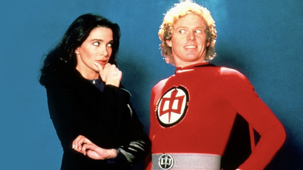 A man in a red superhero outfit stands with a woman in front of a blue screen in The Greatest American Hero