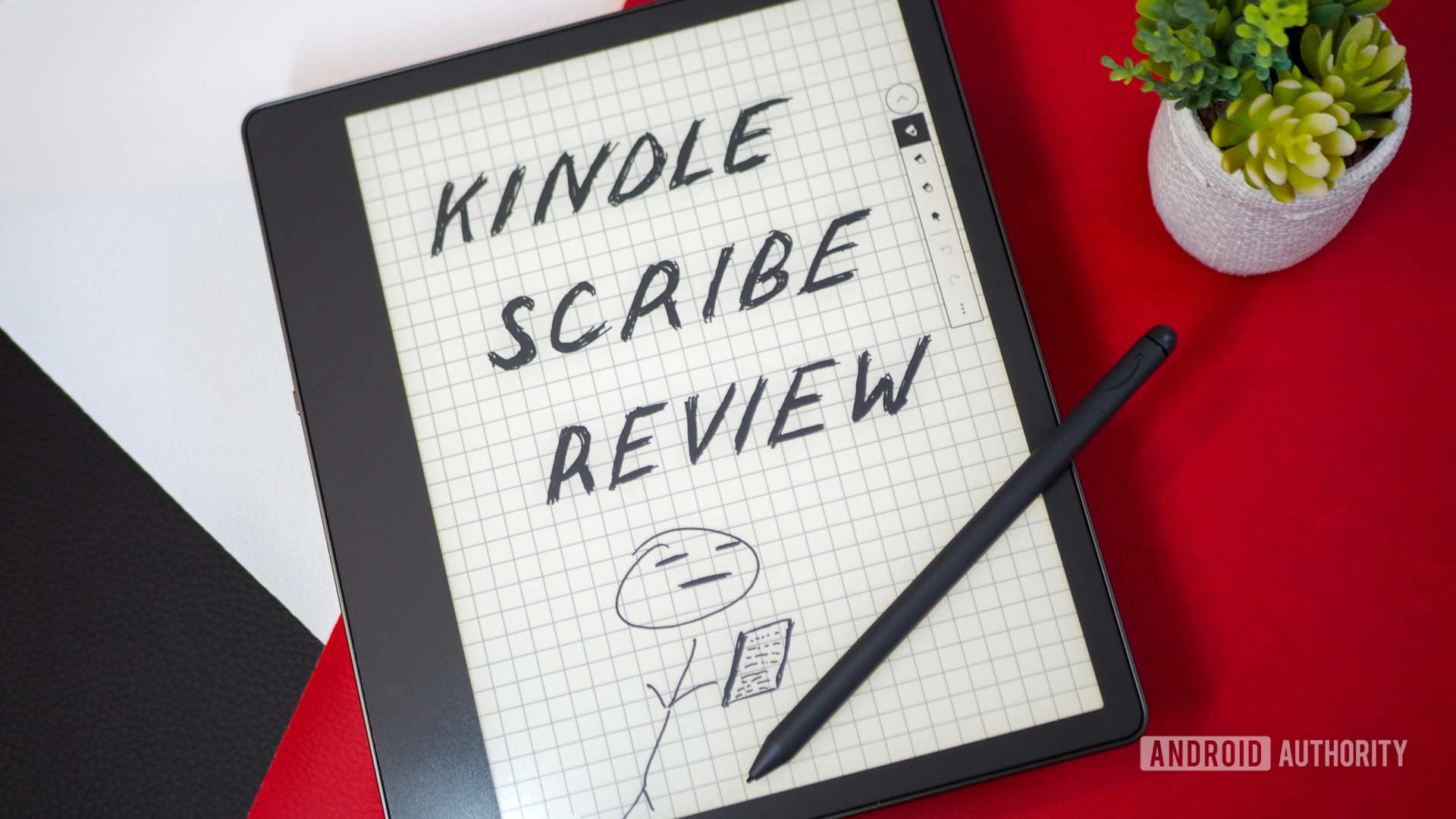 Amazon Kindle Scribe showing a sketch of "Kindle Scribe Review" and stickman holding an e-reader with the Premium Pen next to it