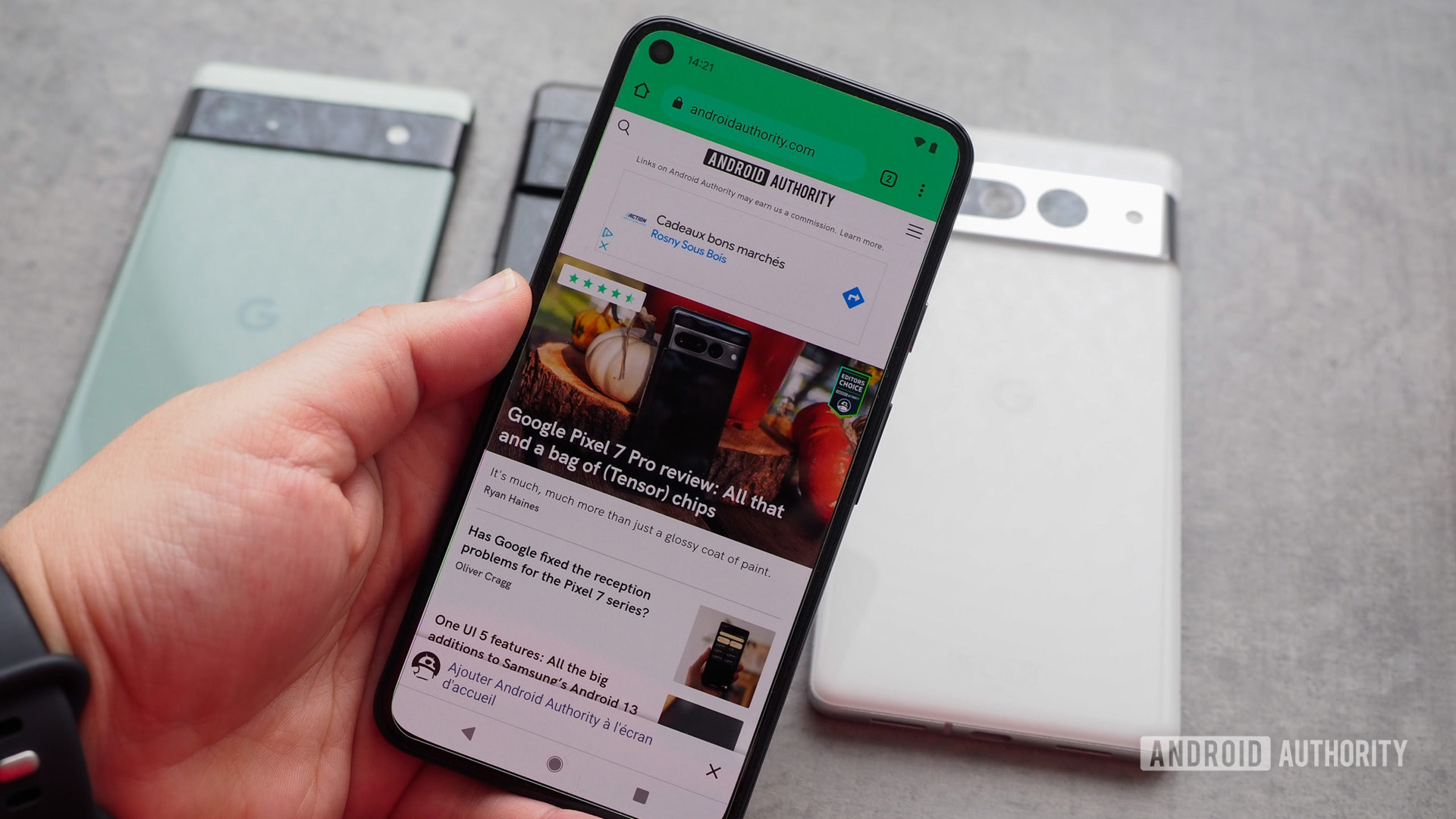 Google Pixel 5 di tangan dengan tampilan menyala, menampilkan situs web Android Authority, dengan beberapa Pixel di belakangnya di atas meja