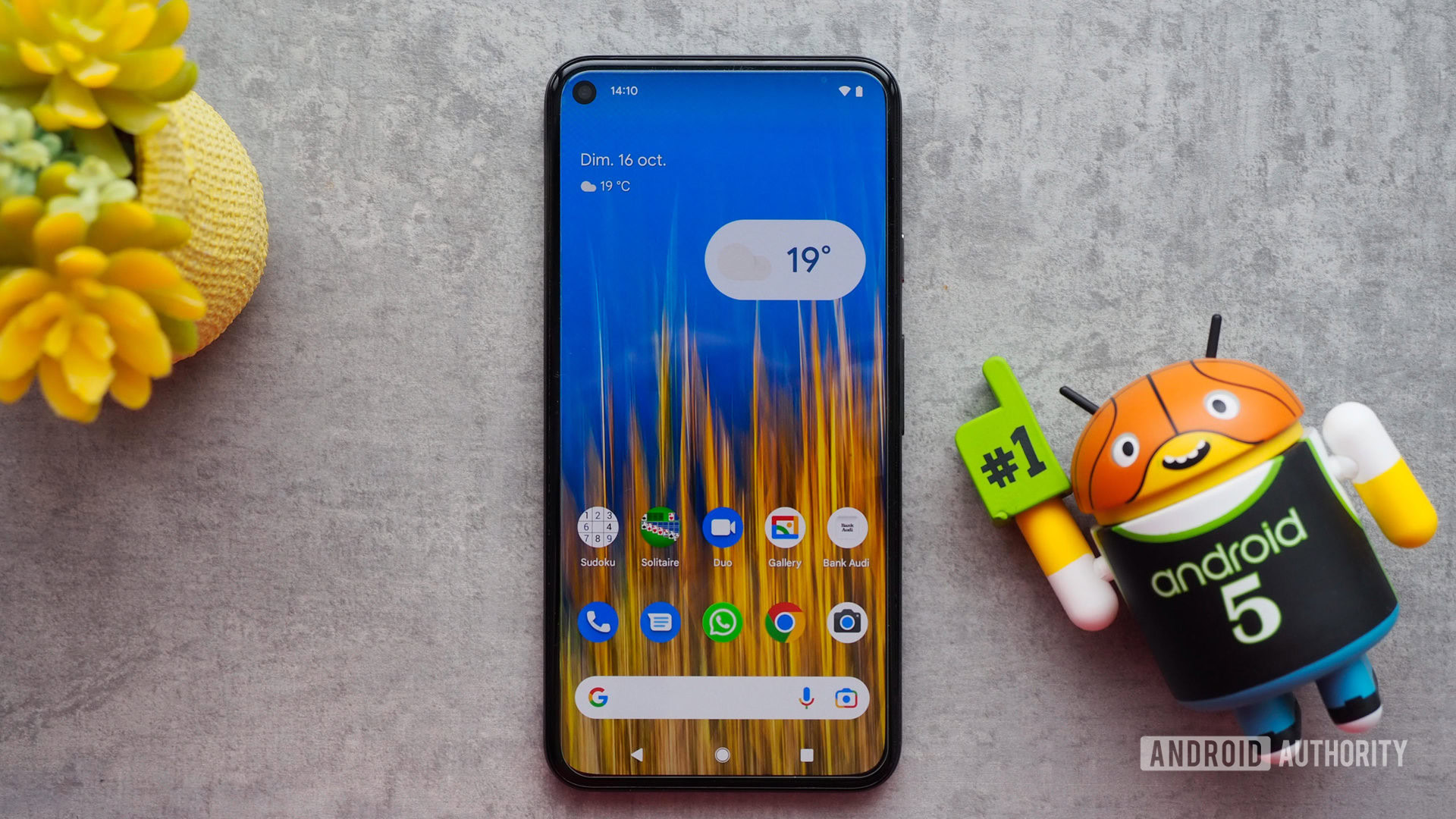 Google Pixel 5 seen from the front with the display on, showing the homescreen, laying on a table with a mini Android basketball figurine next to it