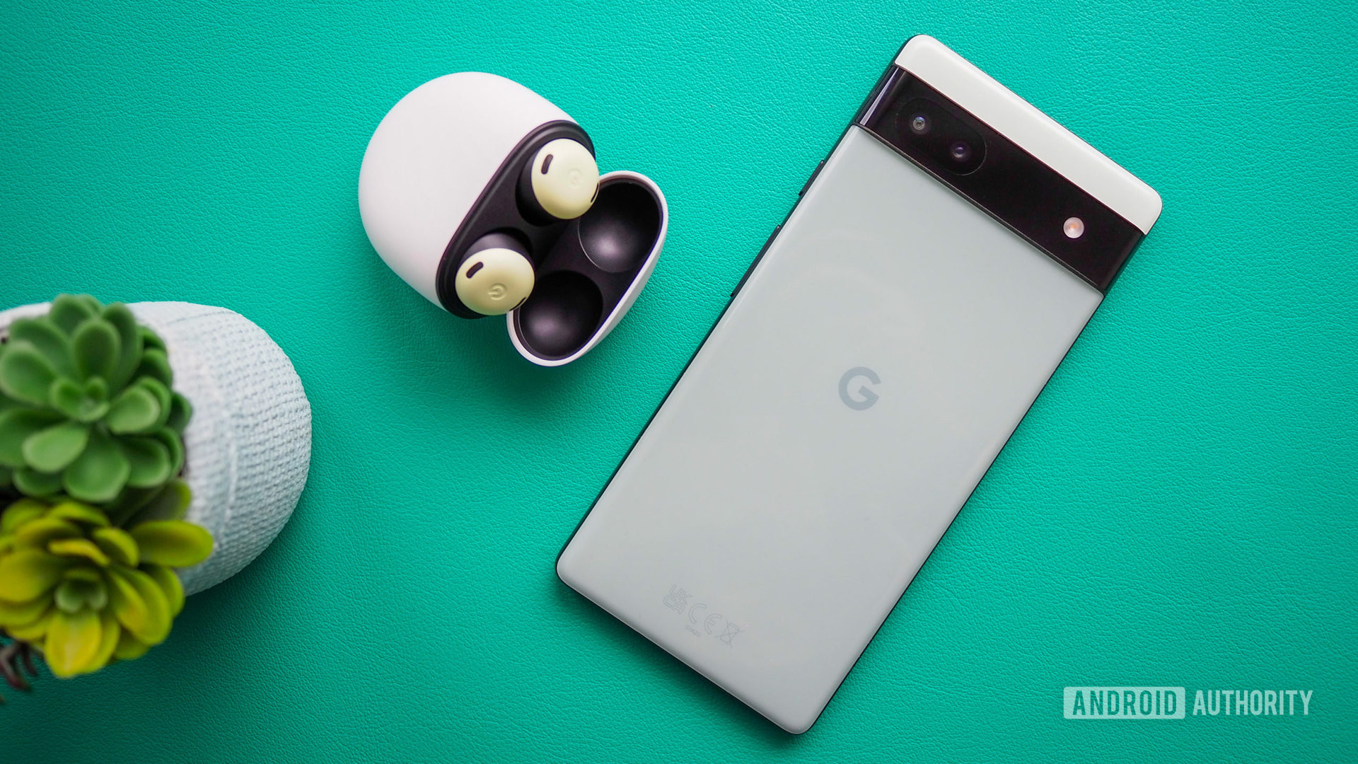 Google Pixel 6a in Sage color, seen from the back, next to Pixel Buds Pro, on a turquoise background