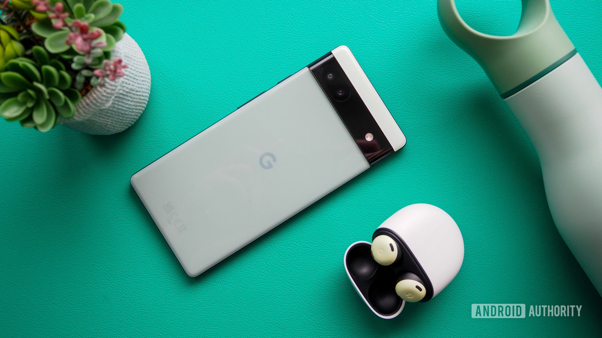 Google Pixel 6a in Sage color, seen from the back, next to Pixel Buds Pro, on a turquoise background