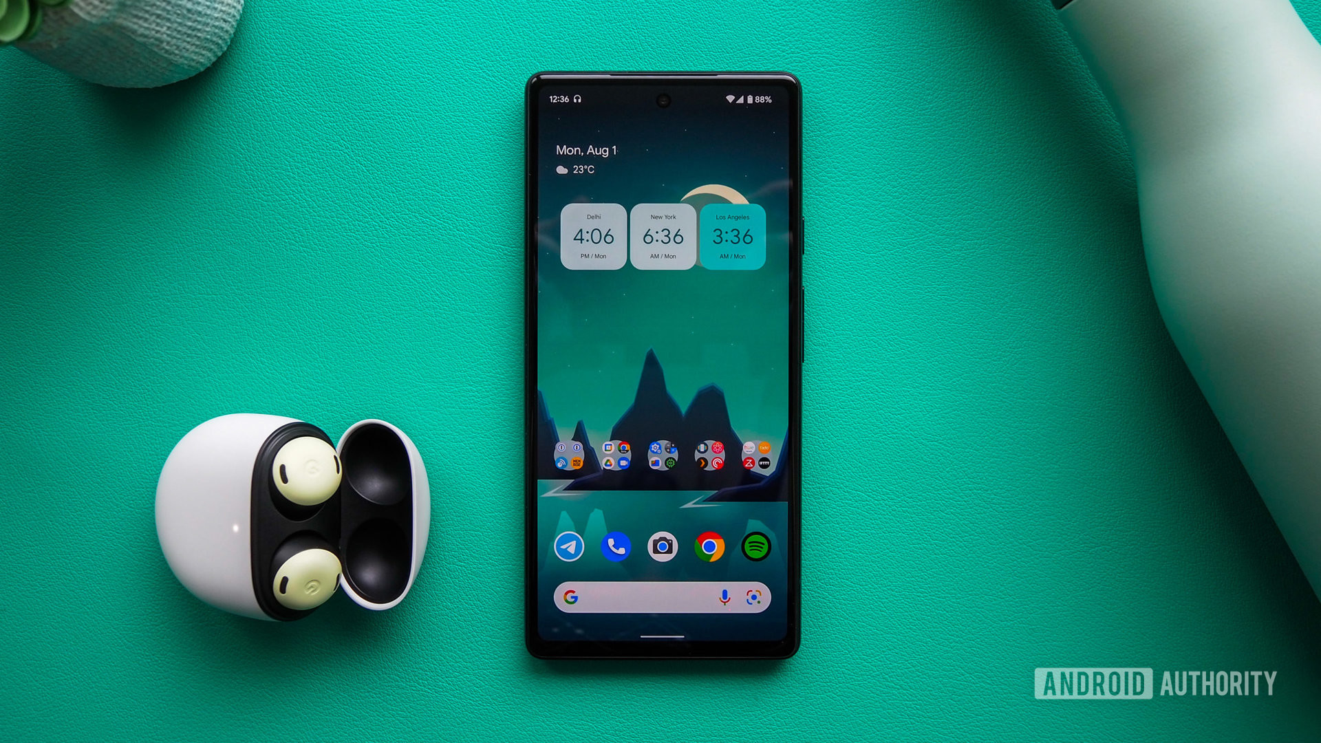 Google Pixel 6a, display on with the homescreen showing, next to Pixel Buds Pro, on a turquoise background