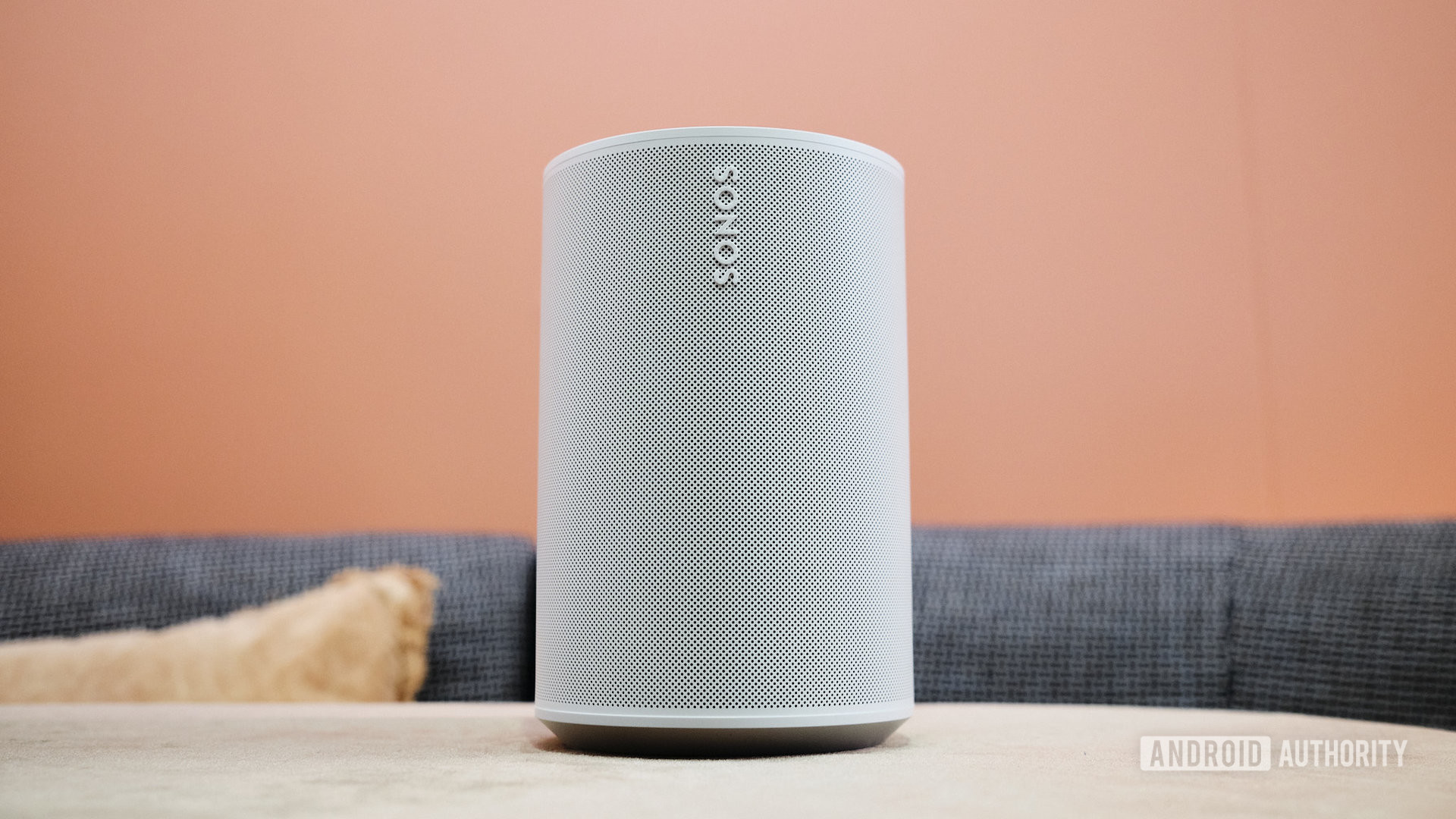 The Sonos Era 100 smart speaker on a table in front of an orange wall.