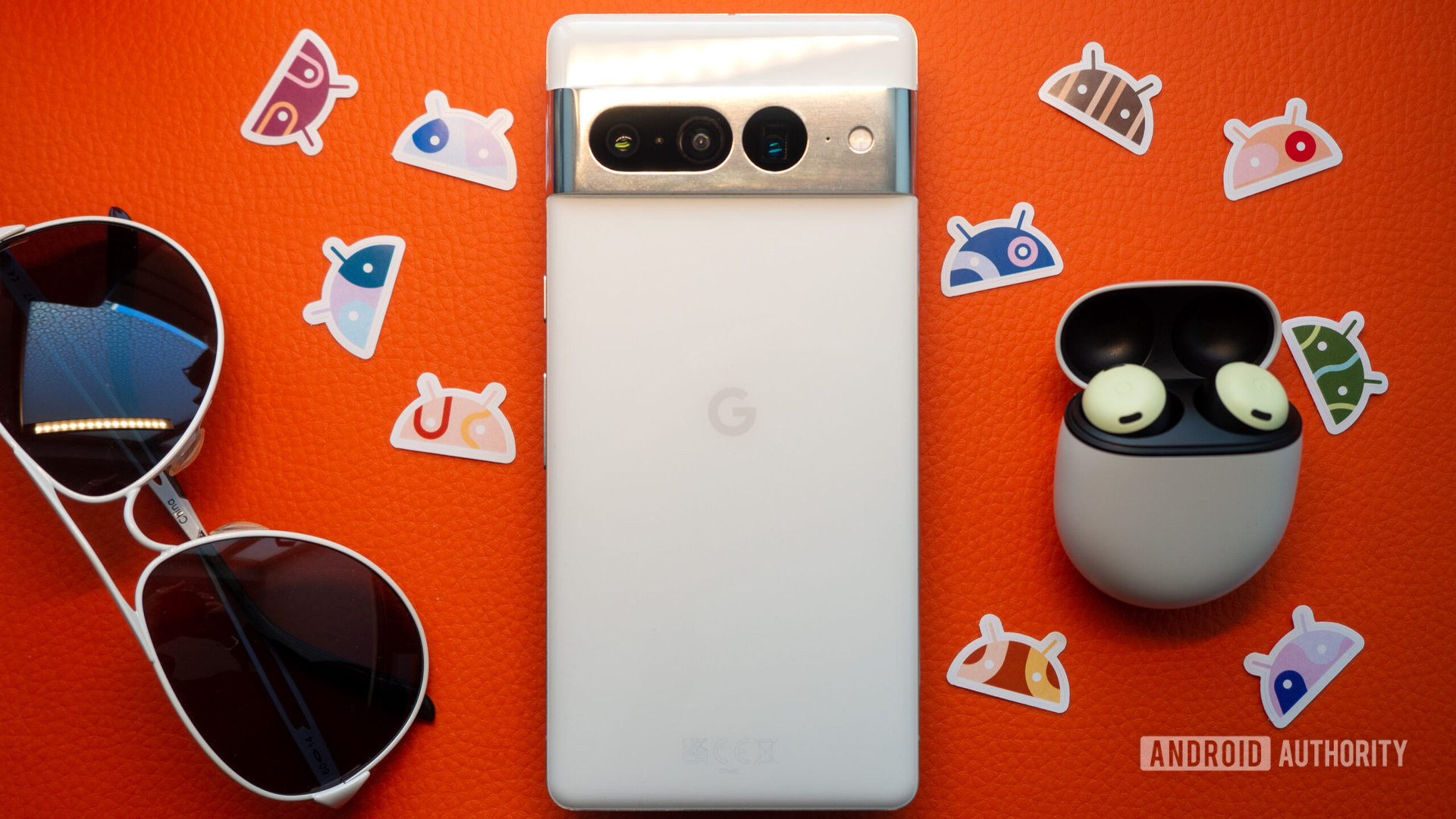 Google Pixel 7 Pro in white and Pixel Buds Pro and white sunglasses on an orange background, with Android stickers next to them