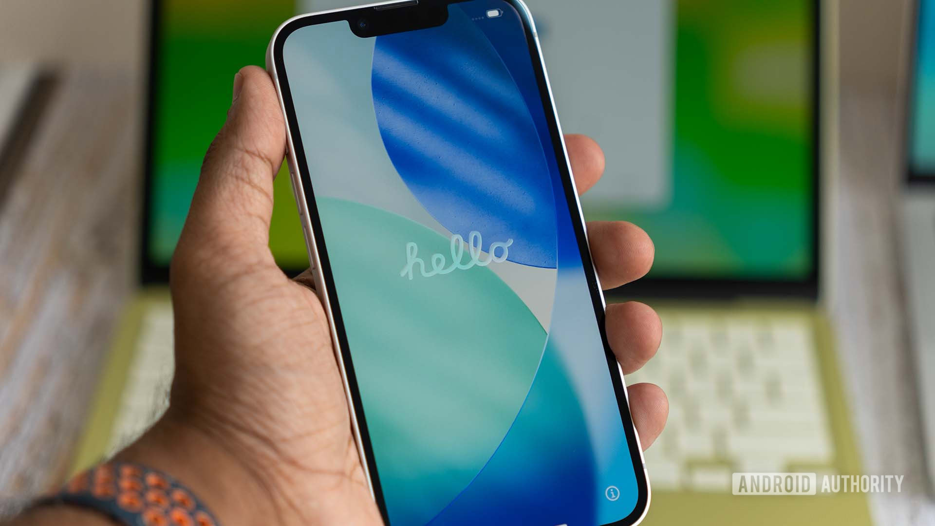 Apple iPhone 17e showing hello startupscreen on display during boot, with MacBook Neo in the background