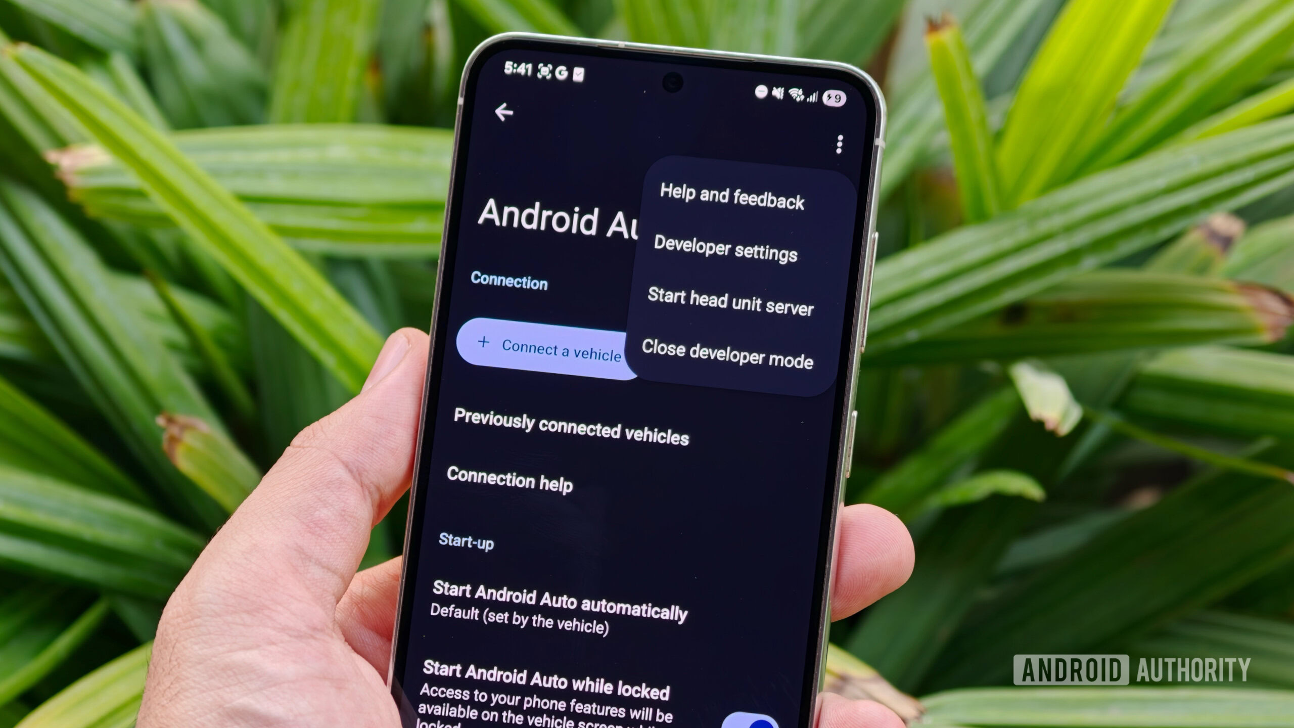 A hand holding a smartphone showcasing the Android Auto settings screen with the Developer settings overflow menu open