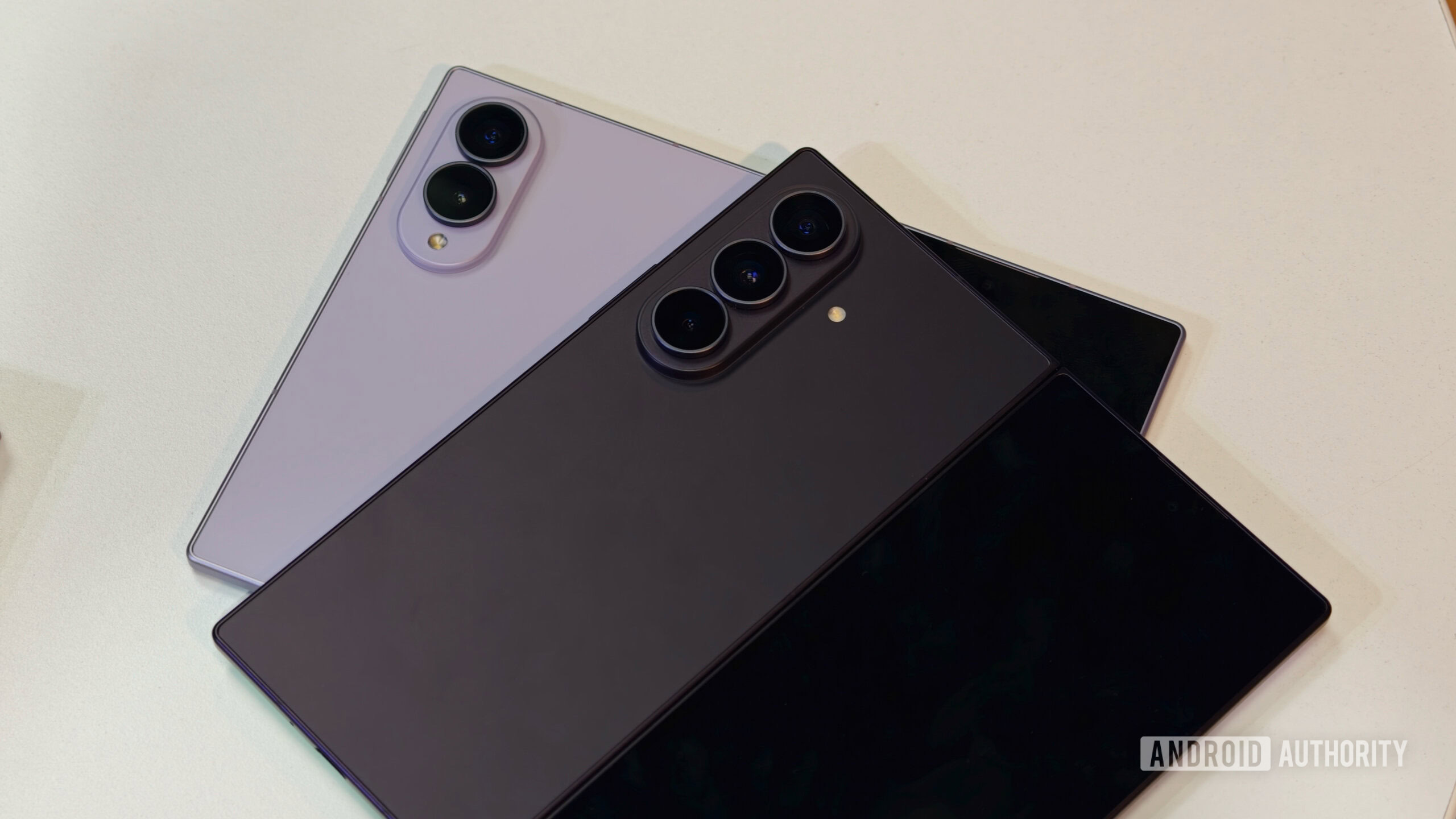 z fold 8 vs ultra 1 Samsung Galaxy Z Fold 8 in lavender resting beneath the darker Samsung Galaxy Z Fold 8 Ultra on a white surface, highlighting their distinct dual and triple camera modules