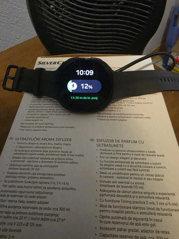 Black Samsung Galaxy Watch 9 resting in front of a desk fan showing 12 percent battery charge at 10:09 AM at the start of a cooling fast charge test.