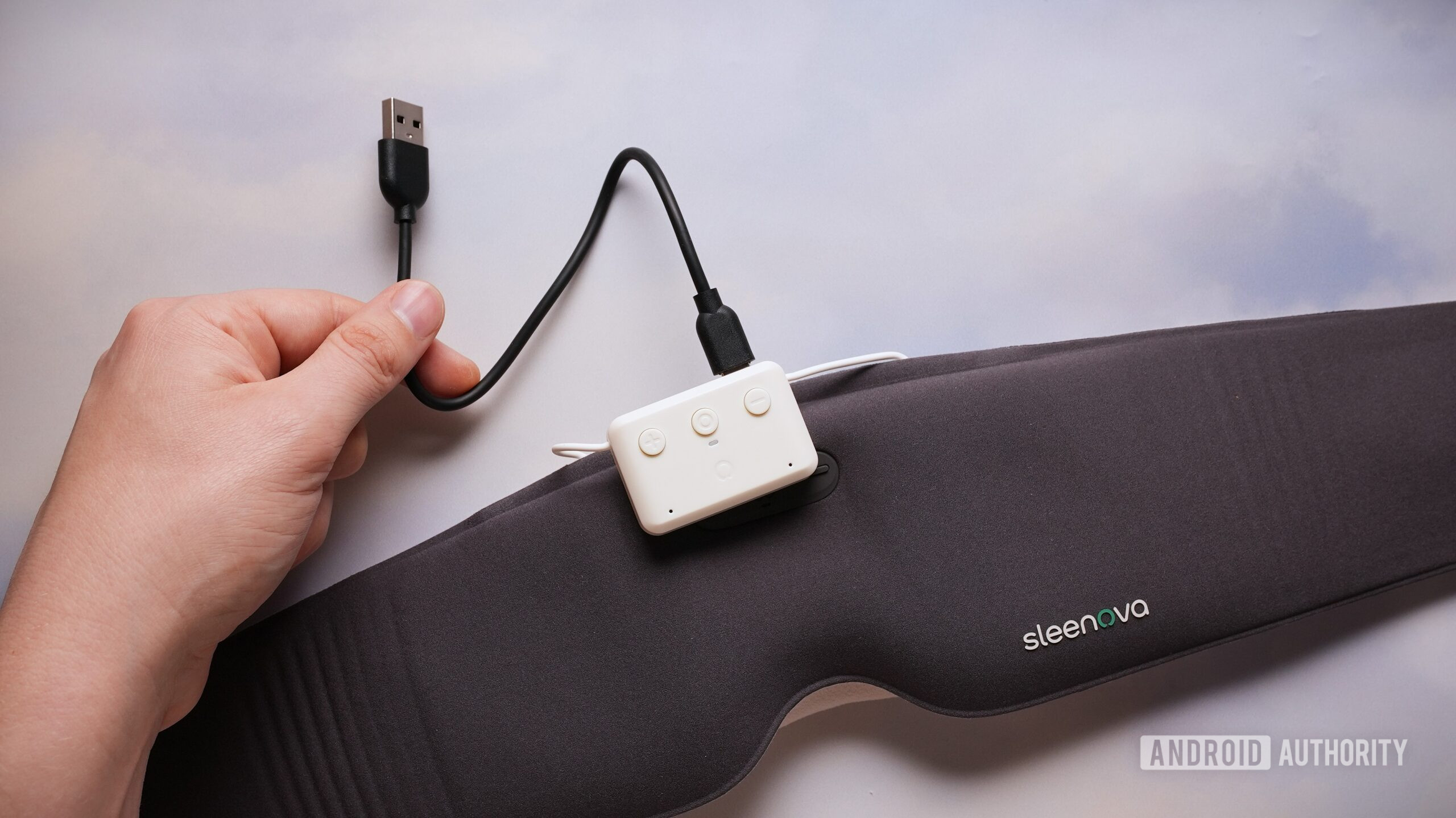 A hand holds the USB charging cable connected to the Sleenova sleep mask’s removable control module.